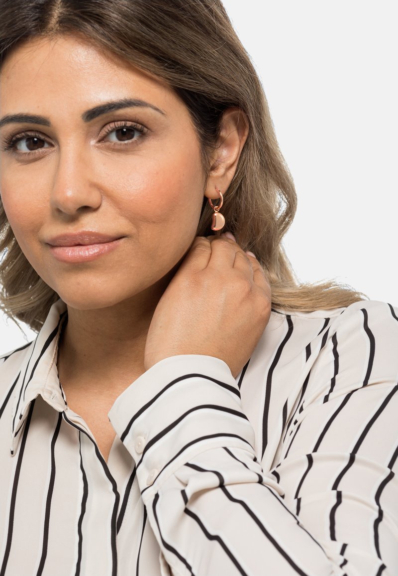 Orecchini a cerchio in oro rosa con un design curvato e semisferico, pendenti dal lobo dell'orecchio, indossati con una camicia bianca a righe nere.