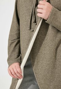 Pullover verde oliva con tessuto strutturato, dotato di una fodera interna bianca e di cordini sul collo, che presenta una vestibilità comoda e maniche lunghe.