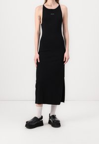 Robe noire sans manches avec un col montant et une fente latérale, fabriquée en matière extensible. Chaussures à plateforme noires au design élégant avec des chaussettes grises.