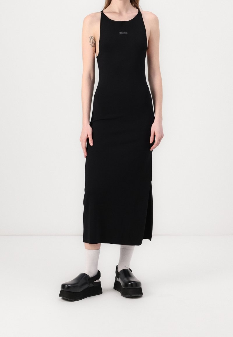 Robe noire sans manches avec un col montant et une fente latérale, fabriquée en matière extensible. Chaussures à plateforme noires au design élégant avec des chaussettes grises.