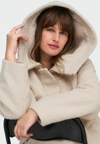 Hele beež kapuutsiga jakk, mis on valmistatud pehmest, tekstureeritud kangast, millel on ees tõmblukk ja lõdva lõikega varrukad, asetatud juhuslikult toolile.