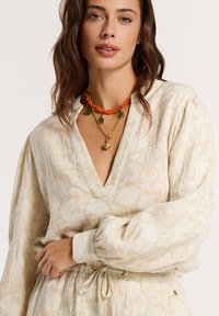 Blusa color crema con un motivo floreale, scollatura a V, maniche lunghe e vita cintrata. Accentuata con una collana a strati arancione e oro.