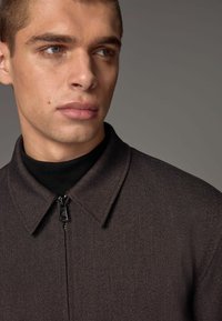 Dark, textured jacket with a collar and zip closure, worn over a black turtleneck. Features a subtle pattern and smooth fabric.