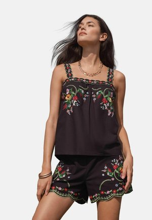 Woman wearing black sleeveless top and shorts with colorful floral embroidery and scalloped edges, accessorized with gold necklace and bracelet.