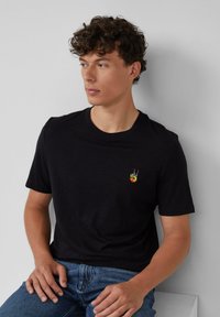 Jonge man met krullend haar, gekleed in een zwart t-shirt met een klein kleurrijk vredesteken patch, zittend tegen een lichtgrijze muur.