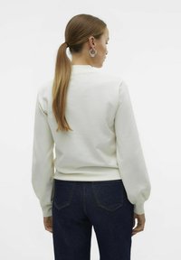 Sweatshirt beige clair avec de longues manches froncées et des poignets côtelés, associé à un jean bleu foncé. Texture lisse, design simple.