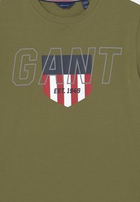 Olive zielona koszulka z dużym szarym napisem "GANT" nad wzorem tarczy z niebieskimi, czerwonymi i białymi paskami oraz napisem "EST 1949" na dole.