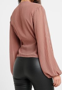 Texturovaný hnědý top s dlouhým rukávem, který má vzor houndstooth a vypasovaný pas, zkombinovaný s černými kalhotami s vysokým pasem.