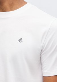 Κοντινό πλάνο ενός ατόμου που φοράει ένα απλό λευκό μπλουζάκι με κοντά μανίκια και μικρά μαύρα γράμματα "M O' P" στο στήθος.