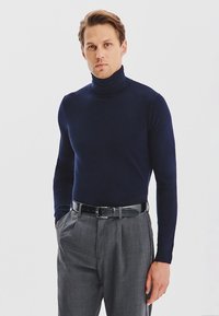 Granatowy sweter z wysokim kołnierzem, wykonany z gładkiej tkaniny, dopasowany krój, w połączeniu z jasnoszarymi spodniami i czarnym paskiem z srebrną klamrą.