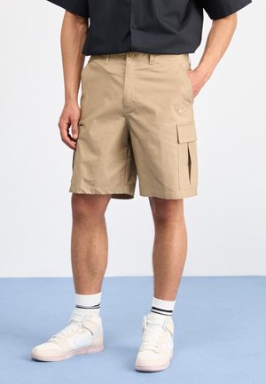 Homme portant un short cargo beige avec des poches, des baskets blanches et des chaussettes blanches avec des rayures noires, se tenant sur un sol bleu contre un fond blanc.