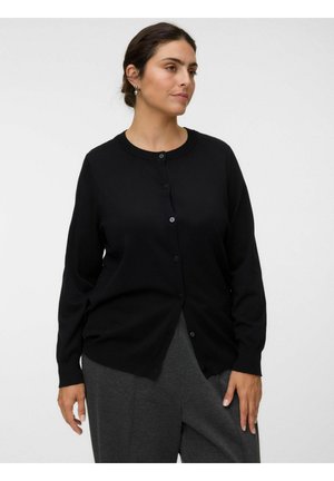 Frau mit dunklem Haar, die eine schwarze, geknöpfte Strickjacke und graue Hose trägt und vor einem einfarbigen hellen Hintergrund steht.