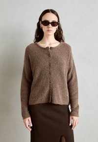 Gilet en tricot marron avec un col rond, fermeture à boutons, manches longues et un ourlet légèrement court. Associé à une jupe marron côtelée.