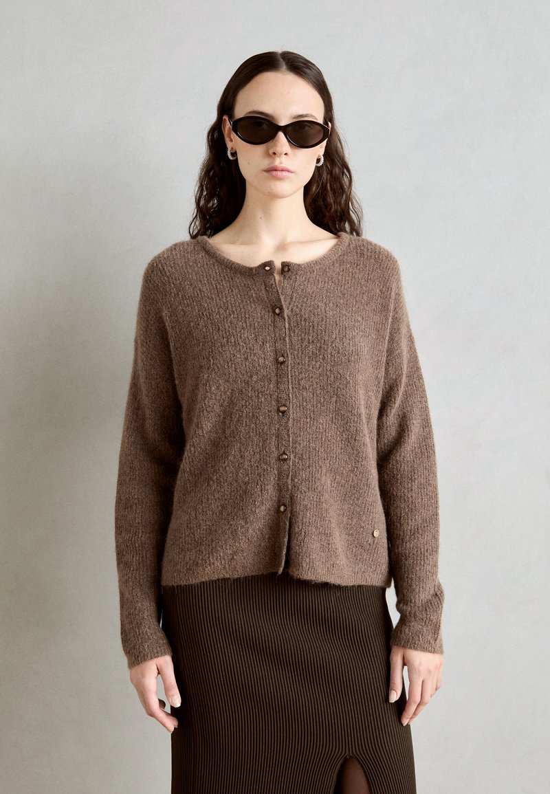 Gilet en tricot marron avec un col rond, fermeture à boutons, manches longues et un ourlet légèrement court. Associé à une jupe marron côtelée.