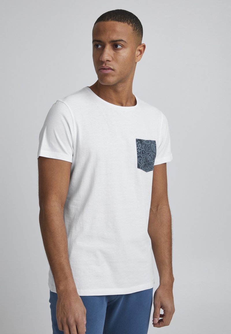 Camiseta blanca de manga corta hecha de algodón, con un bolsillo de patrón en navy en el lado izquierdo. Diseño simple de cuello redondo.