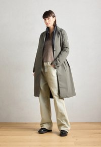 Olijfgroene trenchcoat met een gestructureerd ontwerp, lichtbruine gebreide trui en wijdvallende lichtdenim broeken. Zwarte chunky schoenen maken de look af.