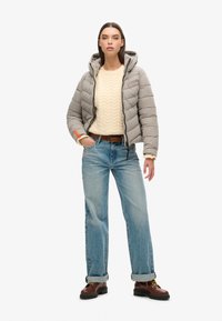 Veste grise rembourrée avec capuche, pull crème en tricot torsadé, jean bleu clair et bottes en cuir marron à lacets ; tenue décontractée en couches avec des détails texturés.