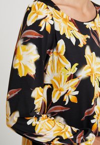 Blusa floral con fondo negro, que presenta grandes flores amarillas y blancas, mangas largas y un escote redondeado. Tejido suave y liso.