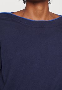 Gros plan sur une personne portant un pull marine avec un large col bordé de bleu, laissant apparaître le cou et le haut de la poitrine.