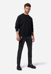 Schwarzer Sweatshirt mit gerippten Bündchen, kombiniert mit eng anliegenden schwarzen Lederhosen mit glatter Textur. Das Modell trägt schwarze Knöchelstiefel.