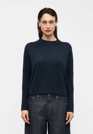 Kvinde med mørkt hår iført en marineblå langærmet sweater og løse mørke denimbukser, stående mod en ensartet lys baggrund.