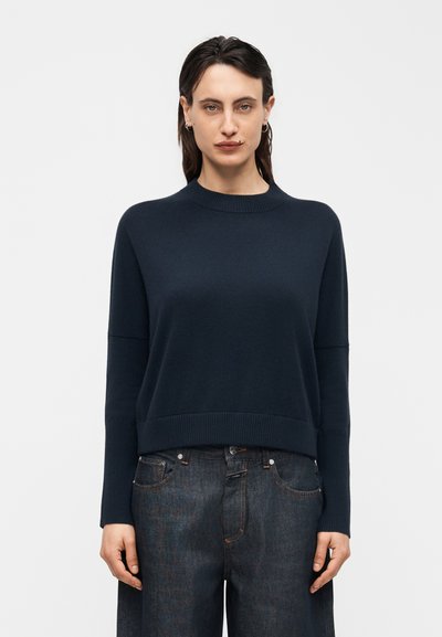Femme aux cheveux foncés portant un pull bleu marine à manches longues et un jean denim foncé ample, debout devant un fond clair uni.