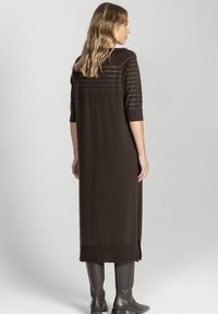 Kobieta ubrana w długą, ciemnobrązową, dzianinową sukienkę z rękawami do łokci i delikatnymi poziomymi paskami, w połączeniu z ciemnymi kozakami za kolano, zwrócona tyłem.