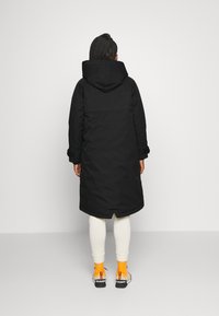 Černá parka s kapucí, vyrobená z hladkého materiálu, v dlouhém siluetu a s elastickými manžetami. V kombinaci s bílými legínami a barevnými botami.