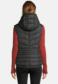 Vrouw met halflang haar die een zwart gevoerd mouwloos vest met capuchon draagt over een rode geribde lange mouw top, met de rug naar de camera gekeerd.
