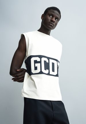 Jaunas vyras dėvi baltą berankovį marškinėlį su ryškiu "GCDS" logotipu ir juodas kelnes, pozavęs prieš paprastą šviesiai pilką foną.