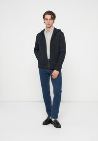 Tumši zils zipa džemperis ar auklu pie kapuces, valkāts pār gaiši pelēku kreklu, kopā ar zilām džinsu biksēm un melnām loafer kurpēm. Gluda tekstūra, ikdienišķa piegriezuma.