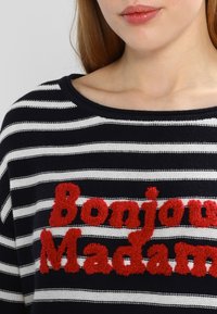 Granatowo-biały sweter w paski z szerokim dekoltem, ozdobiony czerwonym, teksturowanym haftowanym napisem "Bonjour Madame" na przodzie.