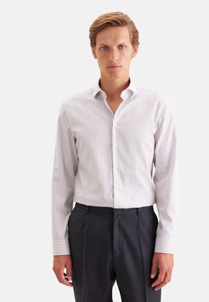 Homme portant une chemise blanche à rayures gris clair avec boutons et un pantalon de costume noir, face à un fond uni.