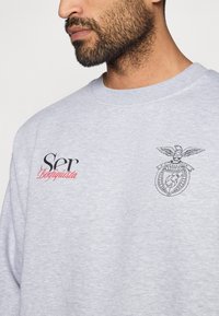 Szürke kapucnis pulóver, kerek nyakkal, texturált anyaggal és hímzett díszítésekkel: "Ser" fekete színben és "Benfiquista" piros színben, egy sas logóval.