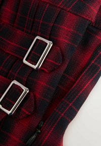 Červeno-černý kostkovaný materiál s kovovými stříbrnými sponami a zipem. Má texturovaný povrch a výrazné vzory.