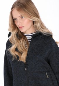 Jachetă ușoară cu model negru și albastru, guler înalt, fermoar frontal și o etichetă pe partea stângă jos. Bluza în dungi dedesubt.