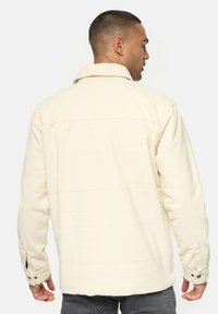 Cremefarbene, strukturierte Jacke mit Kragen, geknöpften Bündchen und horizontalen Nahtdetails auf dem Rücken. Bequeme Passform.