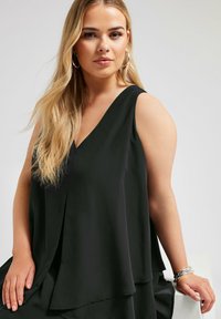 Svart ärmlös topp med V-ringning, lager av tyg, flödande form och mjuk textur, bärs med ett silverarmband på handleden.