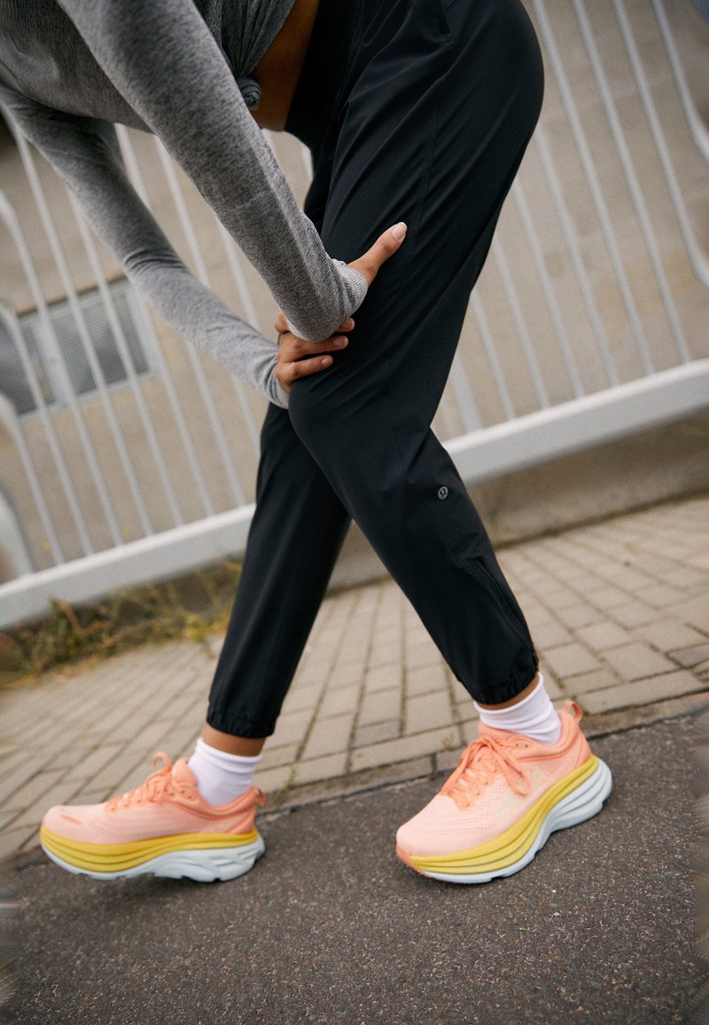Pantofi sport roz și galbeni cu talpă groasă și căptușită, și parte superioară din plasă, asortați cu pantaloni negri tapered și șosete albe până la gleznă.