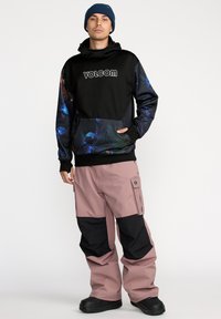 Felpa nera con stampa floreale, dotata di un logo. Abbinata a pantaloni da sci rosa chiaro con accenti neri e numerose tasche. Stivali neri.