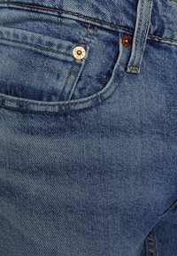 Sötétkék farmernadrág látható varrással, egy elülső zseb fémből készült reteszeléssel, és finom kopásokkal az anyagon.