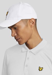 Polo shirt bianco con tre bottoni e un logo di un uccello giallo bordato di nero sul lato sinistro del petto. Un cappellino bianco semplice completa l'outfit.