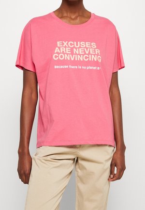 Person iført en pink t-shirt med teksten "UNDskyldninger ER ALDRIG OVERBEVISENDE, fordi der ikke er en planet B" og beige bukser.