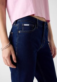 Ciemnoniebieskie dżinsy z kontrastowymi przeszyciami, pięcioma kieszeniami i metalowym guzikiem. Nosi się je z jasnoróżowym krótki topem o luźnym kroju.