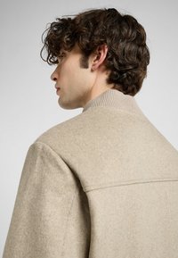 Jonge man met golvend haar draagt een beige jas, zijwaarts weergegeven, waarbij de kraag en textuur van het kledingstuk tegen een neutrale achtergrond worden benadrukt.