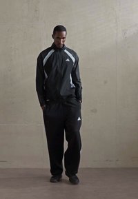 Man in black Adidas tracksuit with white stripes stands against a plain concrete wall with hands in pockets, looking downward.