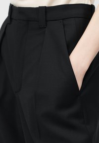 Mustat räätälöidyt housut, joissa on laskokset ja vyölenkit, puoliksi käsi sivutaskussa, yhdistetty vaaleanbeigen yläosan kanssa.