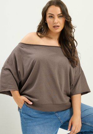 Kvinde med langt brunt hår iført en løs brun off-shoulder top og blå jeans, poserer med den ene hånd i lommen mod en ensfarvet baggrund.