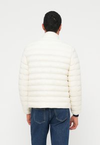 Valge polsterdatud jakk horisontaalsete õmblustega, kõrge kraega ja elastsete varrukaservadega. Kombineeritud siniste teksapükstega, vaadatuna tagant.