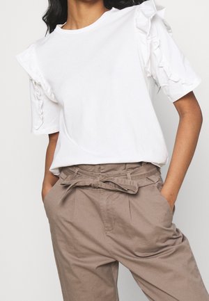 Vrouw die een witte top met korte mouwen en ruches draagt, gestopt in beige high-waisted broek met een ceintuur die geknoopt is. Handen in de zakken.